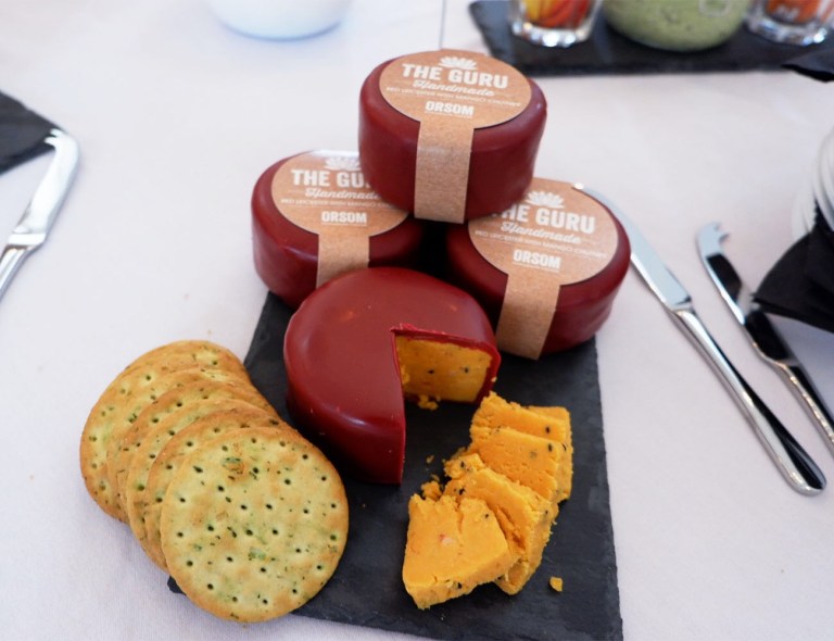 Three Guru cheeses stacked up as a tier the fourth cheese is cut displaying the red leicester cheese from inside, along the left crackers is on display